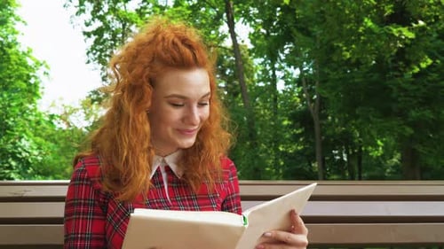 Jovial red haired girl laughing at funny book in park