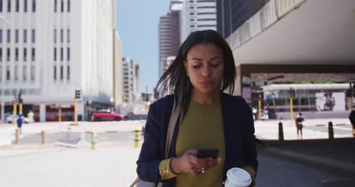 African american woman holding coffee and using smartphone in street