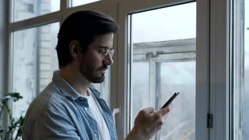 Close-Up Of a man with Glasses Using a Smartphone Online, Standing Near a Window in a Room
