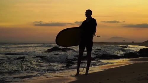 Silhouette eines Mannes mit einem Surfbrett bei Sonnenuntergang, aufgenommen in Zeitlupe.