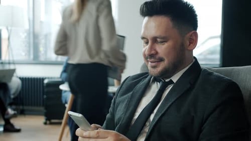Cheerful Businessman Using Smartphone Texting Relaxing in Hotel Lobby with Group of People in Suits