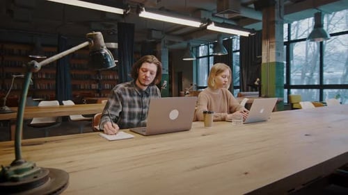 Young Man and Woman Students Studying in Campus Library Researching on Laptops and Taking Notes