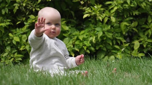 Nettes Baby im Park sitzt auf dem Gras und winkt der Kamera zu