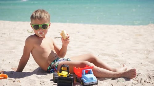 Blonde Boy Eating Ice Cream Sitting on a Sandy Beach