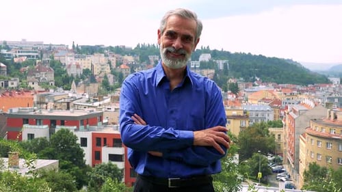 Old Senior Man Smiles To Camera with Folded Arms - City (Buildings) in Background