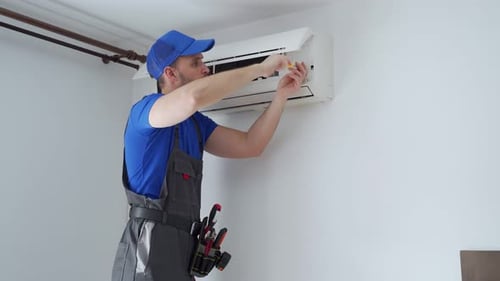 Male Technician in Overalls and a Blue Cap Repairs an Air Conditioner on the Wall