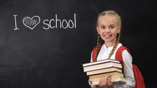 Happy Schoolgirl Holding Books in Front of Chalkboard