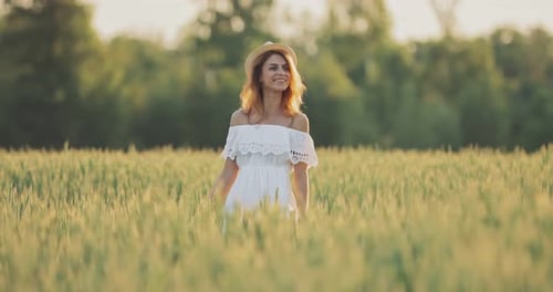 Happy Young Woman in a White Dress and Hat Is Walking in a Green Field. Pretty Girl Walks on a Sunny