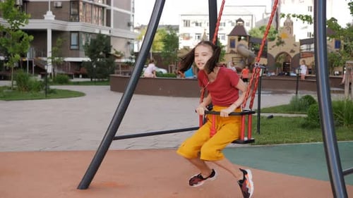 A Girl Rides on a Swing on the Background of a Playground in the Courtyard of a Residential Area
