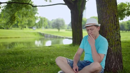 Junger Mann mit Hut sitzt nachdenklich im Gras unter einem Baum