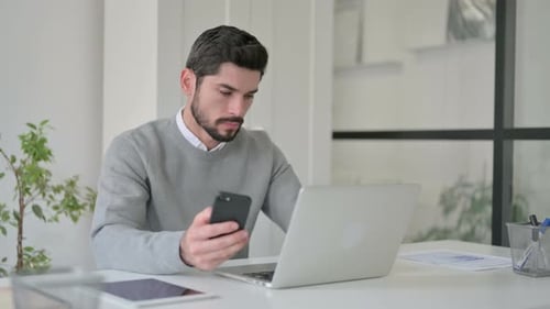 Young Man Using Smartphone While Using Laptop in Office
