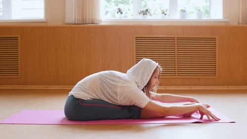Young Slim Woman in White Hoodie Sitting on the Yoga Mat and Leaning Forward