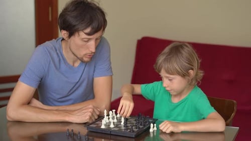 Young Man and His Son Play Chess at Home During the Quarantine. Self-isolation with Kids.