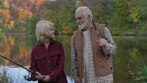 Grandfather with Grandson Smiling and Looking at Camera While Standing Near Lake with Fishing Rods