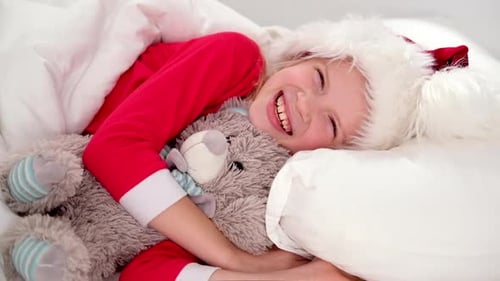 Menina sorridente de chapéu de Papai Noel e pijama de Natal deitada na cama com ursinho de pelúcia em casa