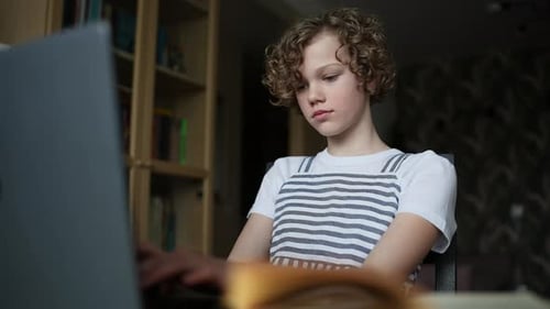 Довольно кудрявая маленькая девочка болтает в социальной сети, печатая сообщение, используя ноутбук, сидящий за столом в