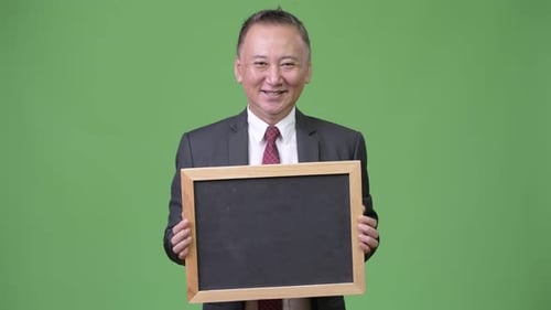 Mature Japanese Businessman Holding Blackboard Against Green Background