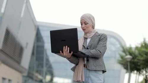 Muslim Woman in Smart Casual Wear Standing Outside Modern Building and Surfing Internet on Laptop