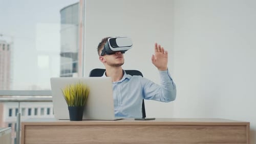 A Young Man Sitting at a Desk in the Office Uses Augmented Reality Glasses To Work on Business