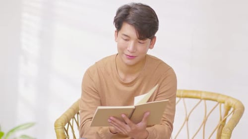 Glücklicher gutaussehender asiatischer Mann sitzt auf der Couch, lächelt und liest ein Buch, entspannen Sie sich zu Hause