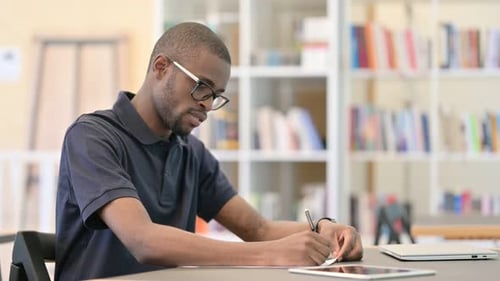 Young African Man Thinking and Doing Paperwork in Library