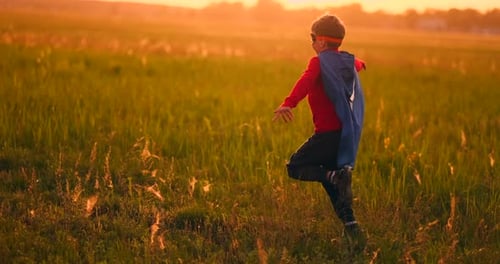 A Boy in a Suit and a Superhero Mask Running Across the Field at Sunset on the Grass