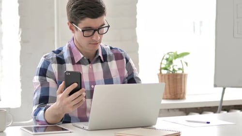 Young Adult Working on Phone and Laptop at Desk
