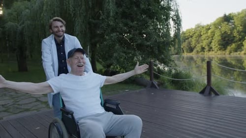 Man Pushing Happy Senior in Wheelchair By Lake