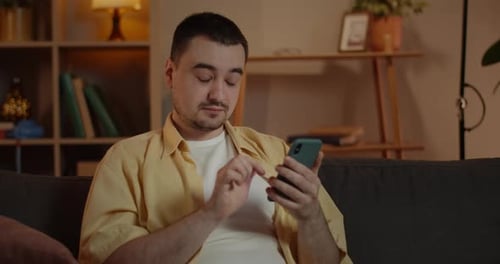 Close Up of Amazed Adult Guy Scrolling and Using Phone Screen While Sitting on Couch in Living Room