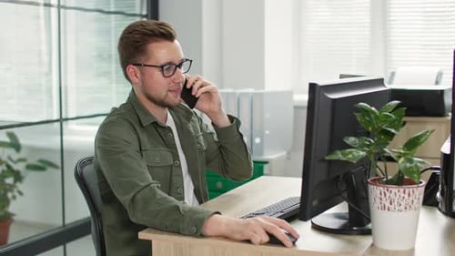 Young Businessman with Glasses Talking on Phone While Working at Computer in Office Background of