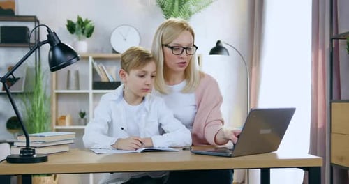 Madre con gafas que se sienta en el escritorio de casa con su apuesto hijo y lo ayuda
