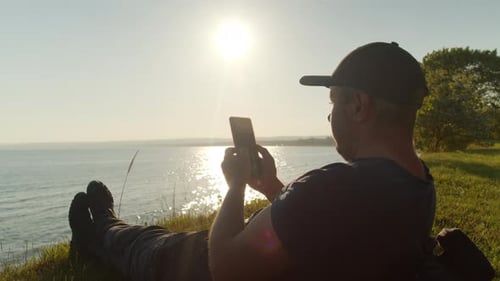 Man Relaxing by Ocean at Sunset with Phone