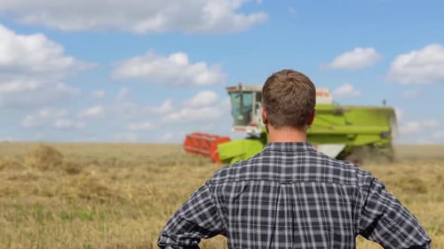 Portrait of the Young Farmer Looking at Combine Working in the Field with Corn and Then Turning His