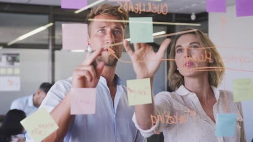 Professional businesswoman and businessman discussing over memo notes on glassboard in modern office