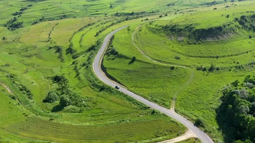 Flying Over a Car Passing on a Countryside Road Winding on a Green Meadow