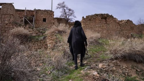 Figure in Black Walks Toward Abandoned Stone Buildings