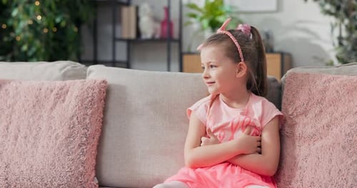 Une petite fille en robe rose avec un bandeau est assise sur un canapé en agitant ses jambes en souriant au repos