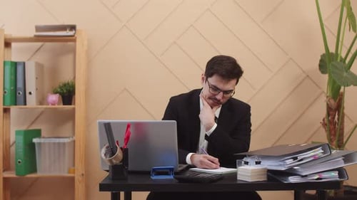 Office Worker Man with Thoughtful Look Writes Something Down in Notebook While Sitting at Table with