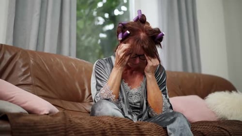 Tired Senior Redhead Woman in Curlers Sitting on Couch Rubbing Temples