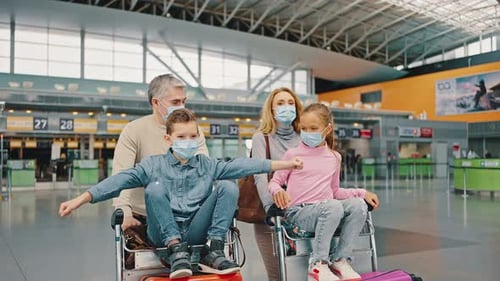 Happy Kids Wearing Protective Masks Riding on Luggage Carts Having Fun at Airport Building Parents