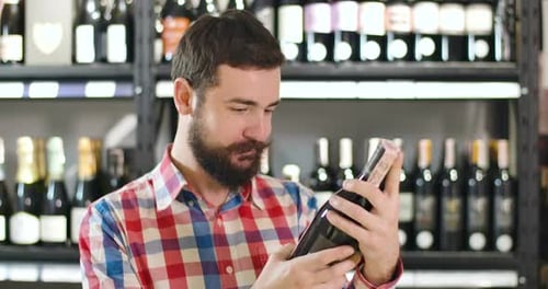 Close-up of Young Confident Caucasian Man Reading Label on Wine Bottle and Looking Back at Shelves