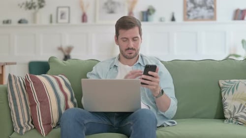 Man Uses Laptop and Smartphone on Couch at Home