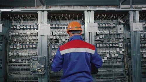 Electrician works at big switch box in helmet and protection jacket. Serviceman is checking voltage