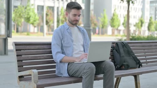 Joven apuntando a la cámara mientras está sentado en el banco al aire libre