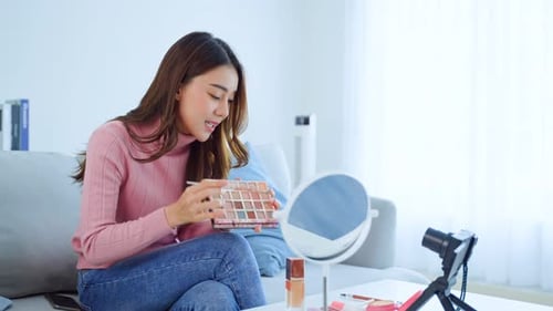 Una vloguera asiática habla con la cámara para revisar el cosmético en casa sentada en el sofá y transmitiendo en vivo.