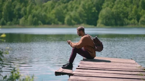 A Young Bearded Man with Glasses and a Backpack Sits on a Wooden Lake Pier in Slow Motion and Prints
