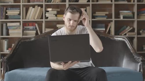 Man Uses Laptop on Couch by Bookshelf