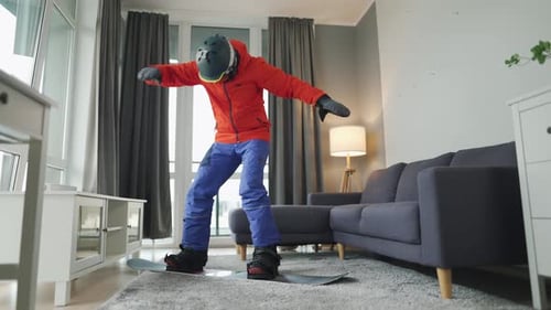 Vídeo divertido. Homem vestido de snowboarder monta uma prancha de snowboard em um tapete em um quarto aconchegante. Esperando por um