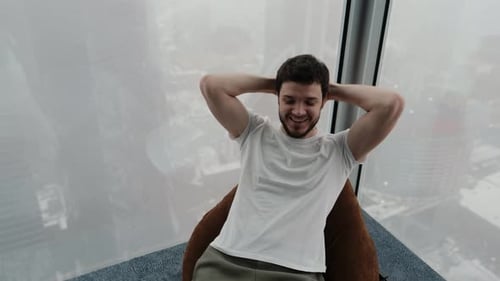 A Young Guy Relaxes on a Pouf in His Modern Apartment Overlooking the City From a Height