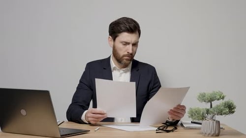 Bearded Man Reviews Documents and Types at Laptop
